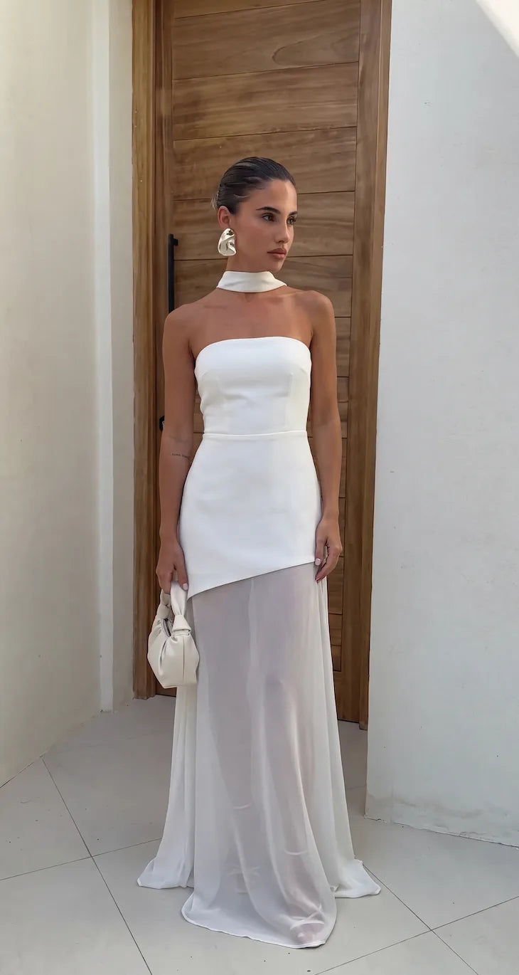 Front view of strapless white asymmetrical maxi dress with attached neck tie and sheer chiffon skirt layer, worn against a timber door