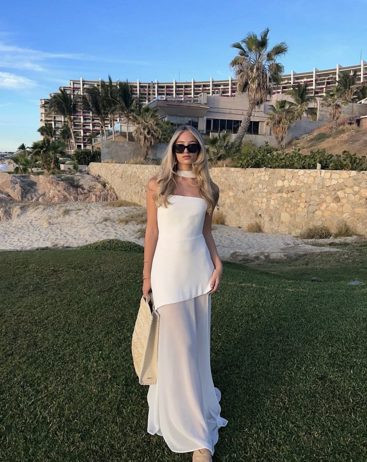 Full-length shot of ivory strapless maxi dress with sheer skirt panel, styled with a straw tote bag and beige sandals on grass