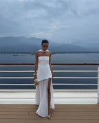 Model wearing white strapless gown with asymmetric high slit and detachable chiffon neck tie, holding a gold top-handle bag on a ship deck
