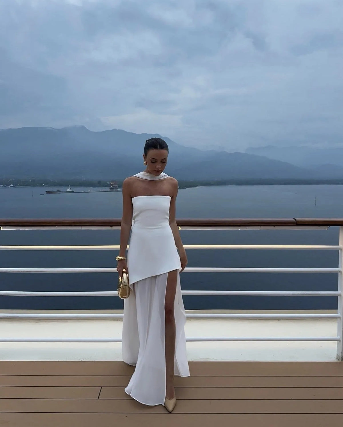 Model wearing white strapless gown with asymmetric high slit and detachable chiffon neck tie, holding a gold top-handle bag on a ship deck
