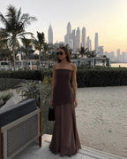 Full-length front view of brown strapless asymmetrical maxi dress with sheer skirt panel, styled with a black crossbody bag on a beach terrace