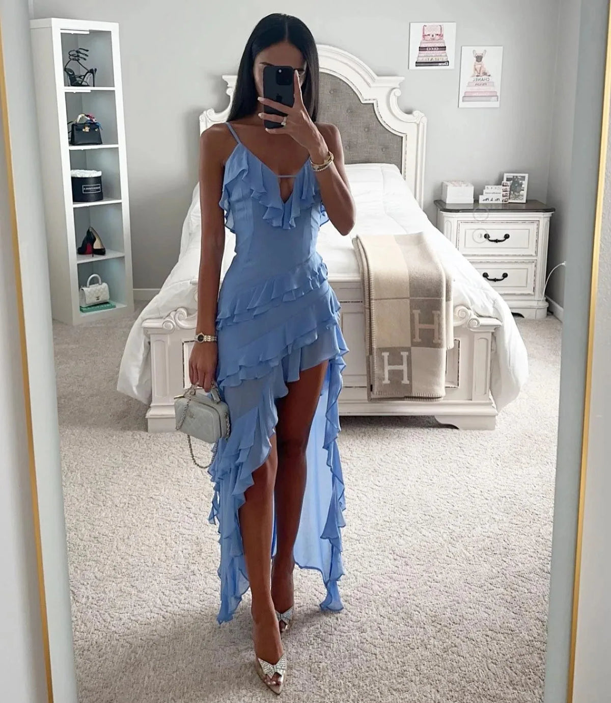 Woman in a sky blue ruffled wrap maxi dress with asymmetric high-low hem, standing in a bedroom with white furniture.