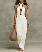 Model wears a white linen halter maxi dress with a criss-cross neckline, paired with a woven bag and beaded necklace
