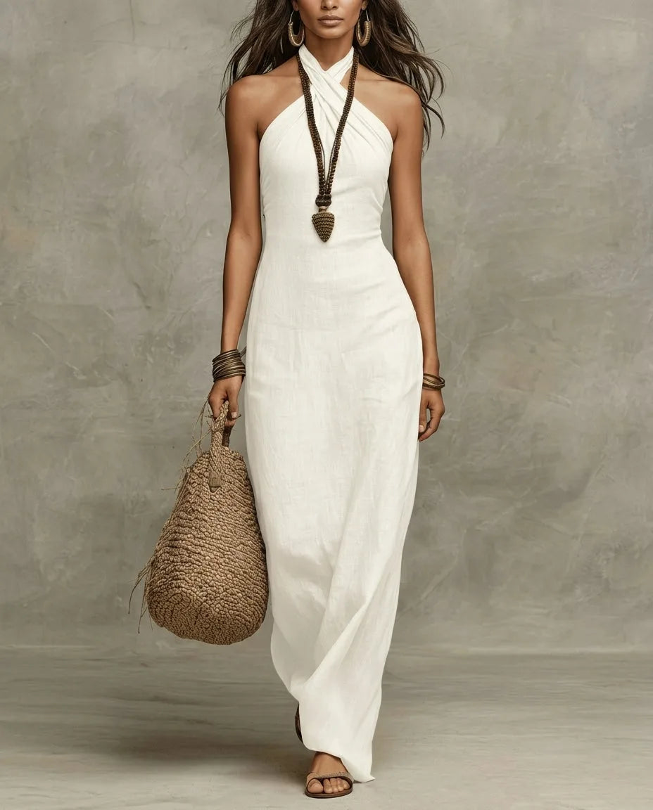 Model wears a white linen halter maxi dress with a criss-cross neckline, paired with a woven bag and beaded necklace