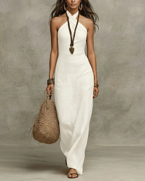 Model wears a white linen halter maxi dress with a criss-cross neckline, paired with a woven bag and beaded necklace