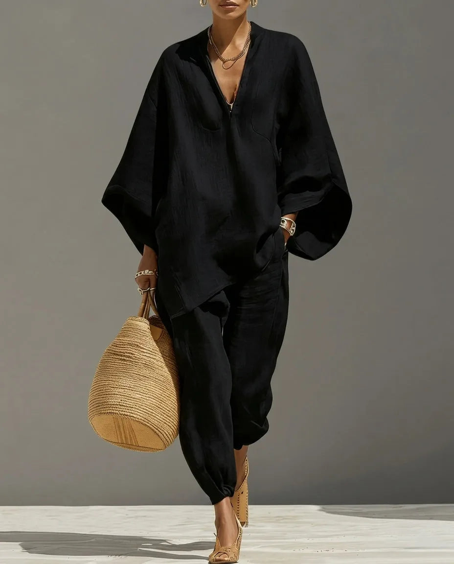 Full-length view of a model walking in a black two-piece set with a V-neck batwing top and tapered pants, styled with a woven tote and heels.