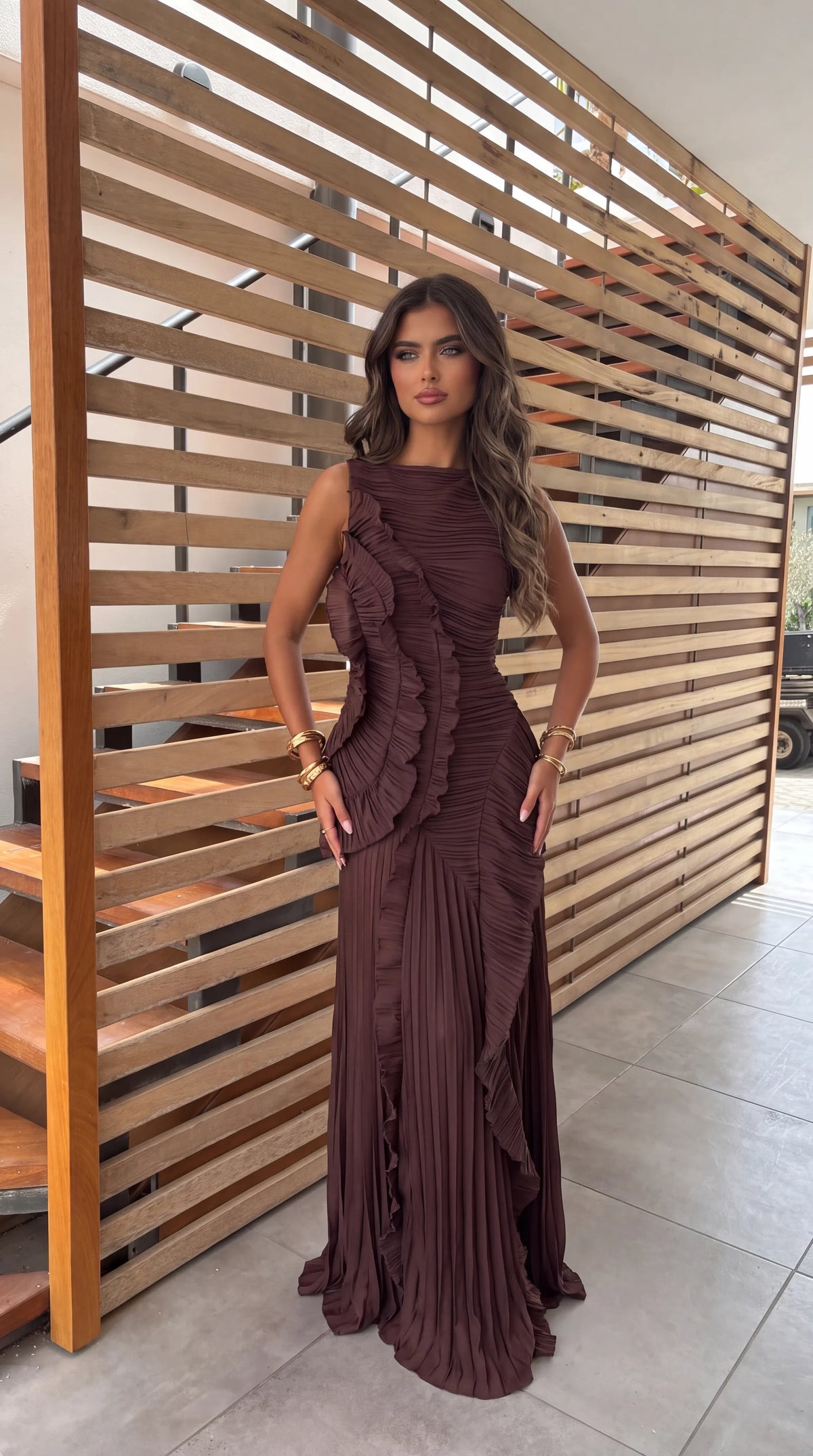 Front view of a model in a brown pleated maxi dress with sculptural ruffle detailing down the bodice and skirt