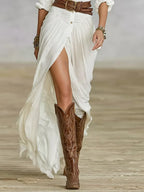 Close-up of the draped white skirt panel with thigh-high slit and button placket, worn with embroidered brown knee-high boots on a wooden floor