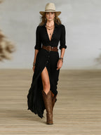 Front view of the maxi shirt dress in black with button placket, thigh-high slit, leather waist belt and brown knee-high boots