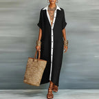 Model wearing a black maxi shirt dress with contrast white placket and collar, holding a straw tote bag