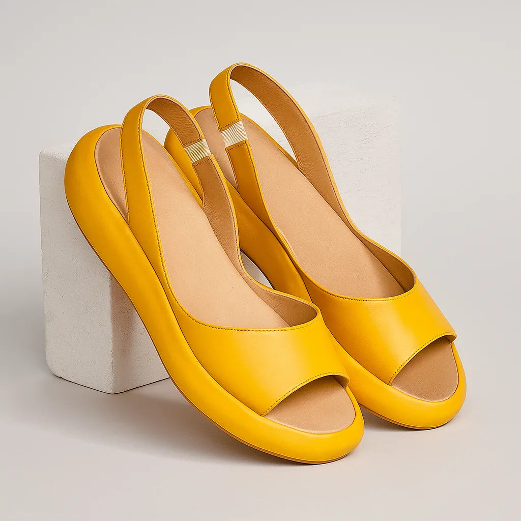 Still-life product shot of a pair of mustard yellow slingback sandals propped against a white block, showing the full silhouette and tan lining.