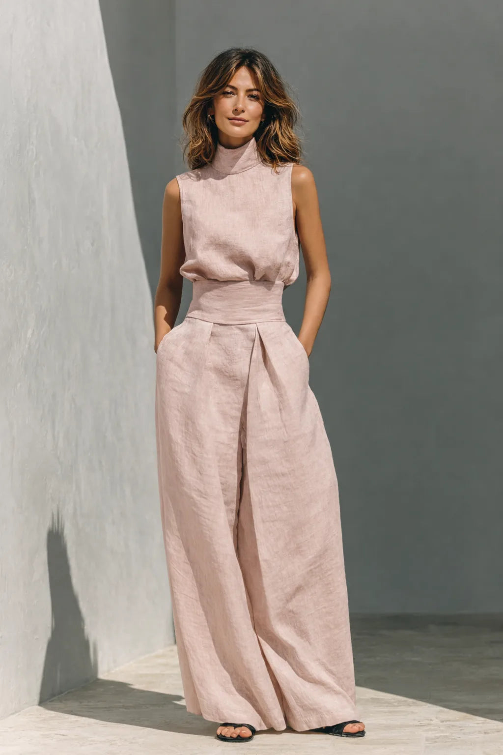 Front view of the sleeveless linen two-piece outfit in a soft light pink shade with wide-leg pleated trousers and black sandals