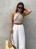 Woman wearing a beige crisscross halter crop top with white textured wide-leg pants, straw tote and gold jewellery