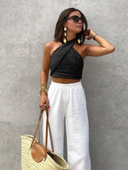 Model wearing the halter top in black, paired with white textured pants and a woven straw tote bag