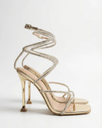 Side view of a gold strappy stiletto sandal with crystal-embellished straps spiralling up the ankle and a square open toe.