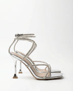 Side profile of a silver mirror-finish stiletto sandal with crystal straps winding around the ankle and a sculpted heel.