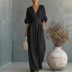 Black deep V-neck jumpsuit with fitted waistband and relaxed wide-leg pants, styled with a woven handbag
