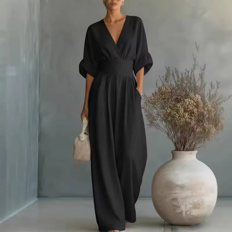 Black deep V-neck jumpsuit with fitted waistband and relaxed wide-leg pants, styled with a woven handbag
