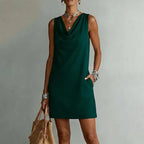 Woman wearing a sleeveless forest green mini dress with a draped cowl neckline and side pockets, paired with layered necklaces