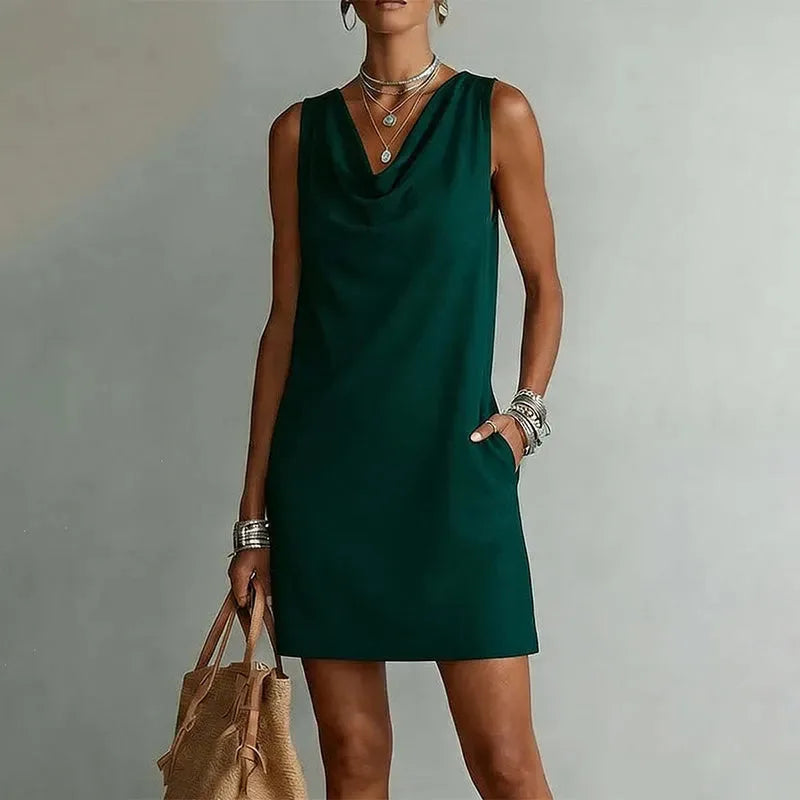 Woman wearing a sleeveless forest green mini dress with a draped cowl neckline and side pockets, paired with layered necklaces
