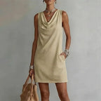 Khaki sleeveless mini dress with a draped cowl neckline, styled with silver bangles and a straw bag
