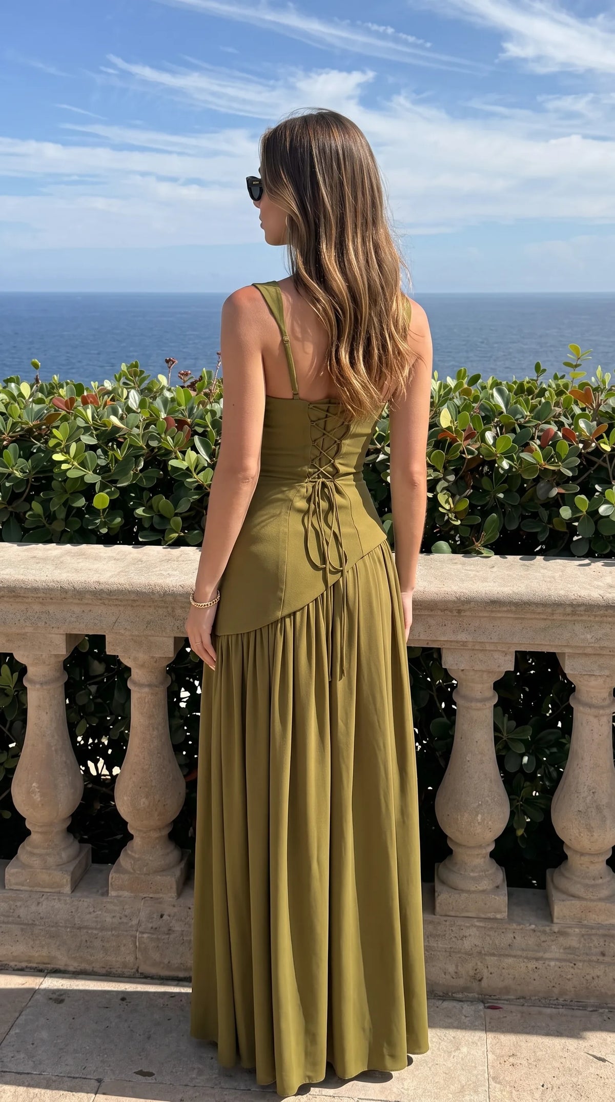 Back view of olive green maxi dress showing lace-up corset back, thin straps and flowing pleated skirt against the sea