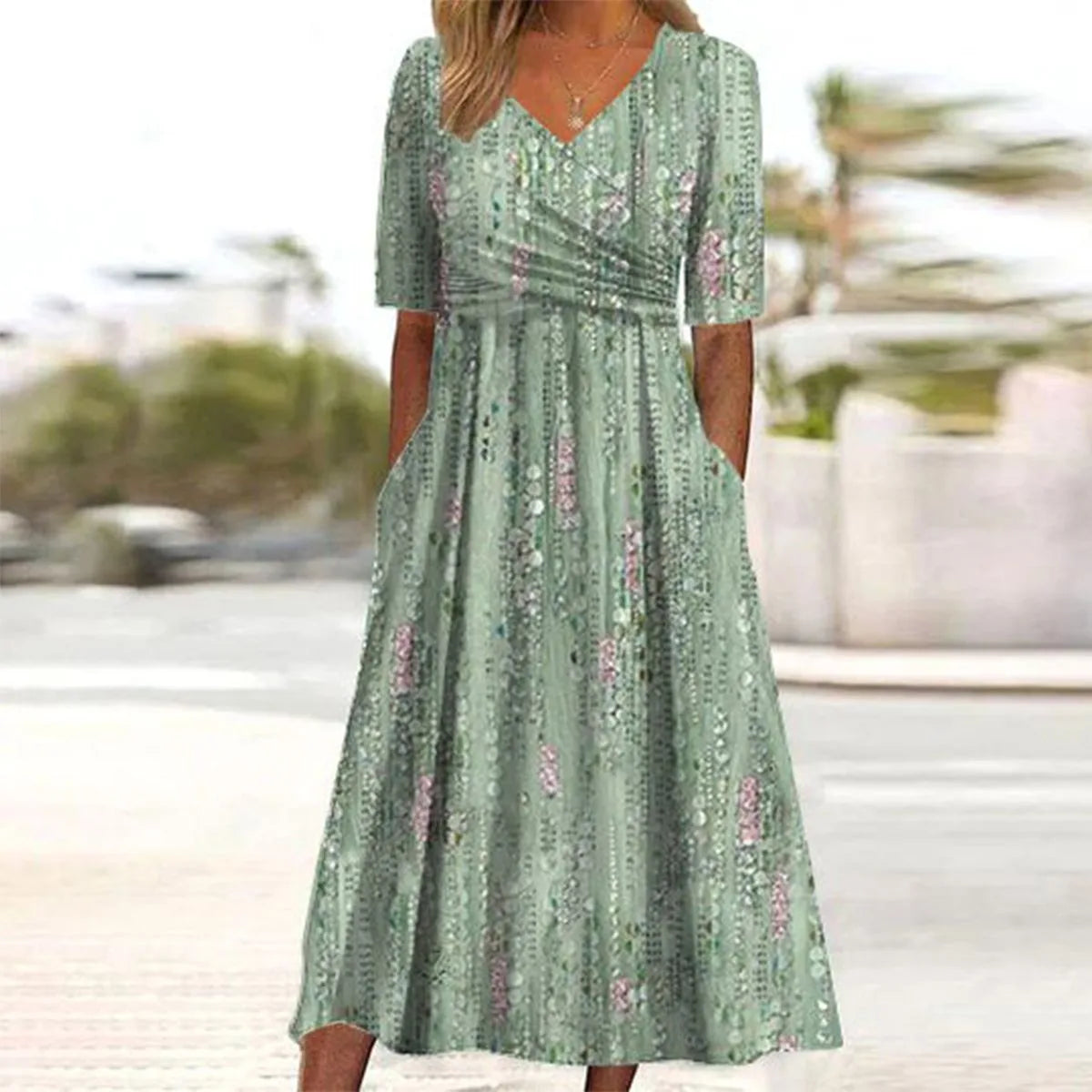 Woman wearing a sage green floral midi dress with wrap-style ruched waist and V-neckline, standing on a coastal path.