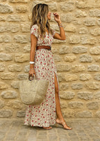 Side view of a woman wearing a beige floral maxi wrap dress with a brown belt and straw tote against a stone wall