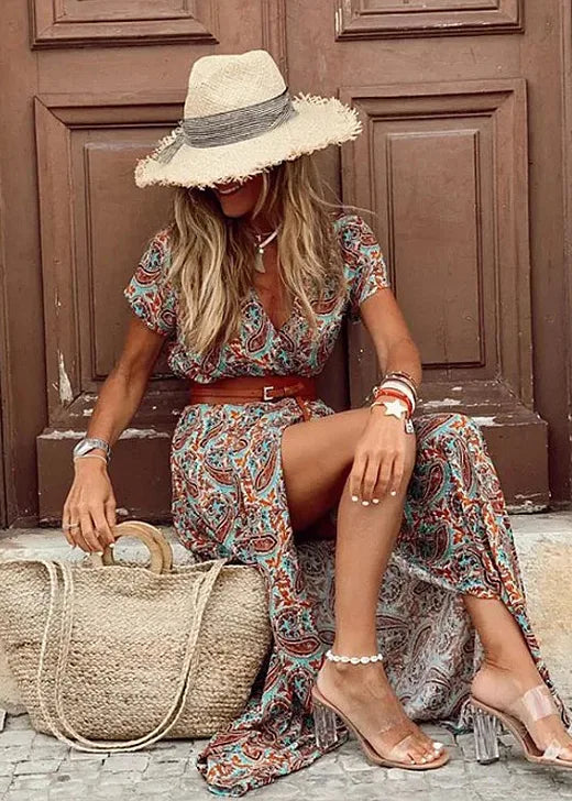 Woman seated wearing a brown and teal paisley maxi dress with a straw hat and leather belt in front of wooden doors