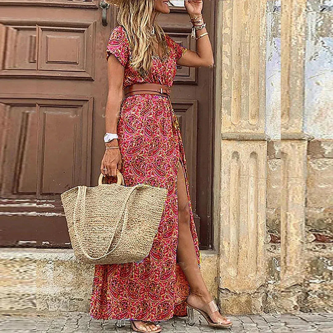 Side view of a woman in a red and pink paisley maxi dress with brown belt, straw tote and heeled sandals