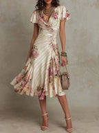 Front view of an ivory satin midi dress with pink floral print, wrap neckline, flutter sleeves and ruched waist, styled with nude heels
