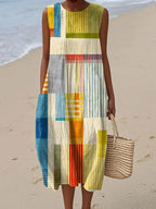 Front view of the sleeveless midi dress in yellow and blue geometric patchwork print, paired with a woven straw tote on the beach.