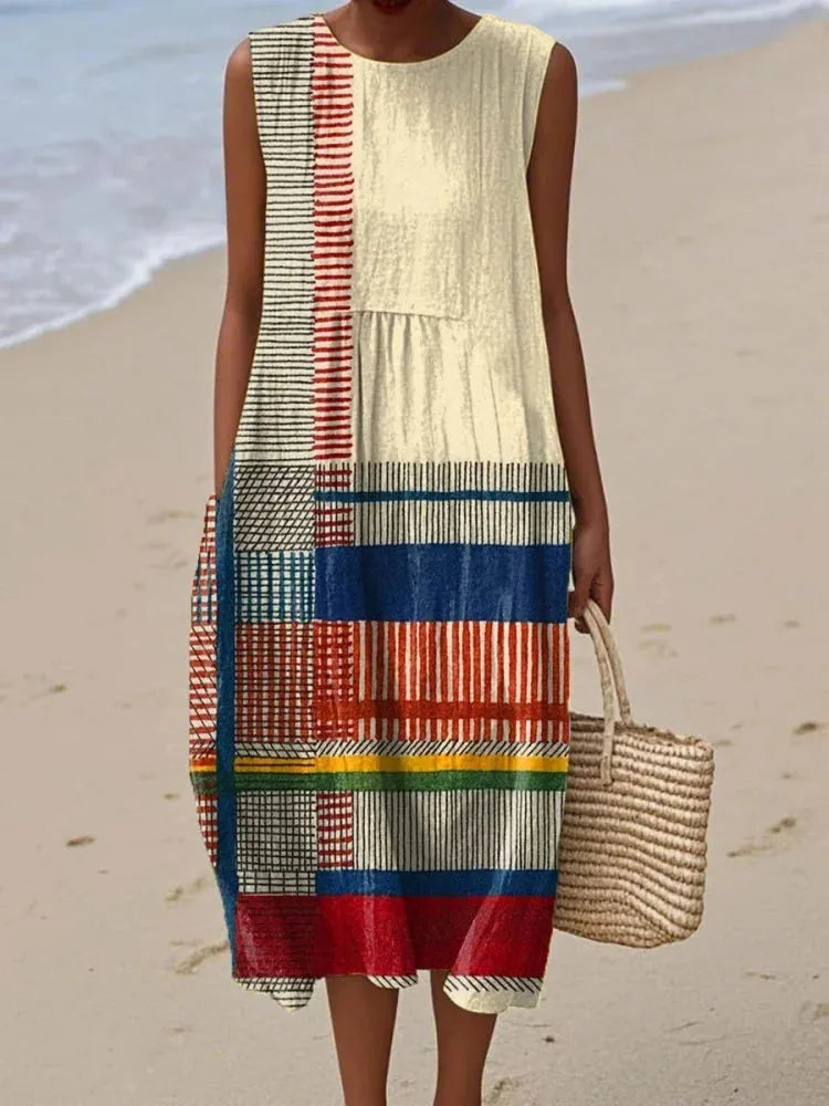 Sleeveless cream midi dress with red, blue and black striped geometric panel detail, worn beside the ocean shoreline.