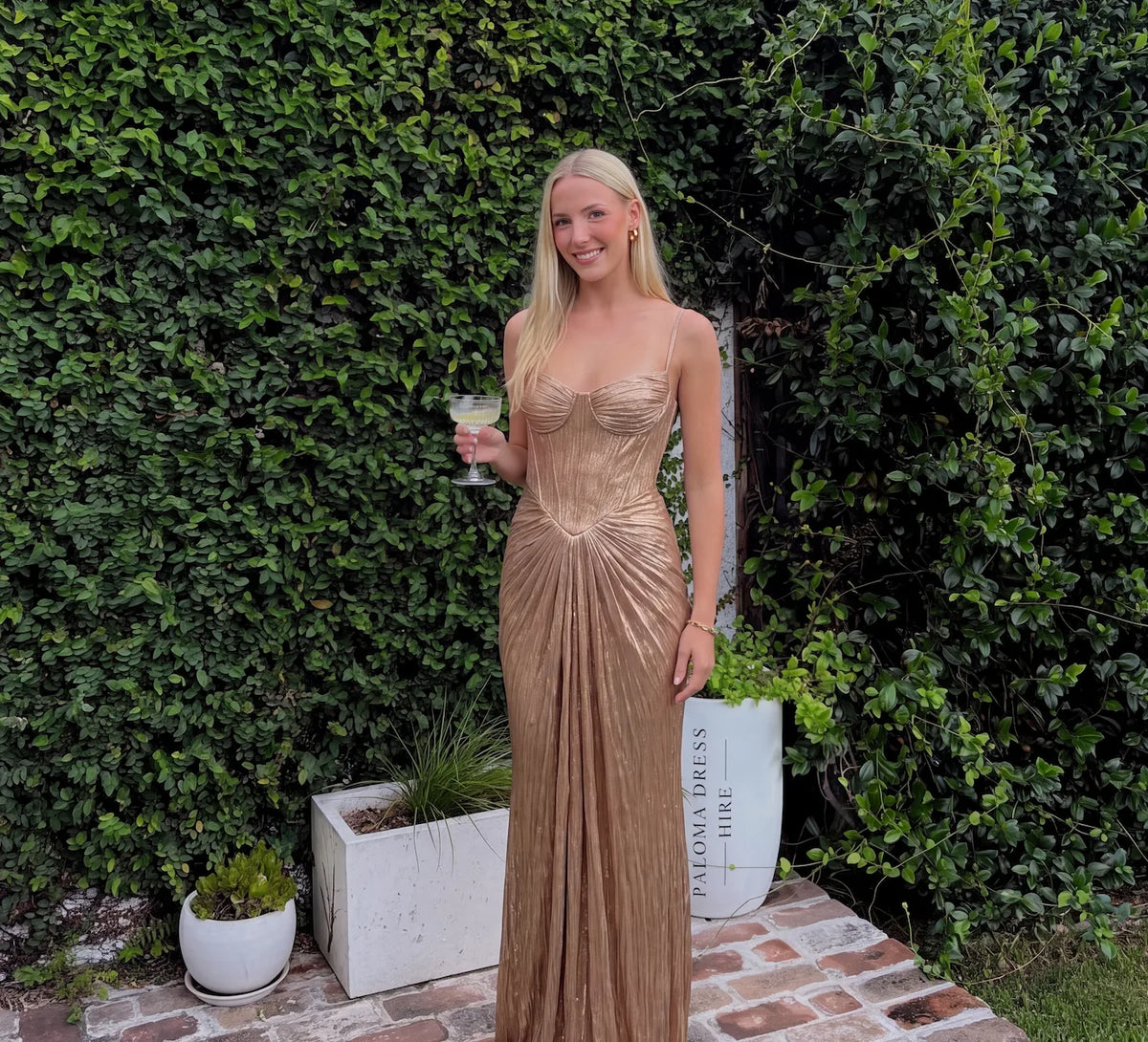Model smiling outdoors in gold shimmer corset maxi dress holding a drink against a green hedge backdrop
