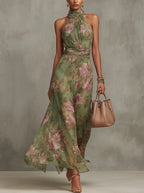 Front view of a woman walking in a green floral halter-neck maxi dress, holding a tan handbag, paired with strappy heels