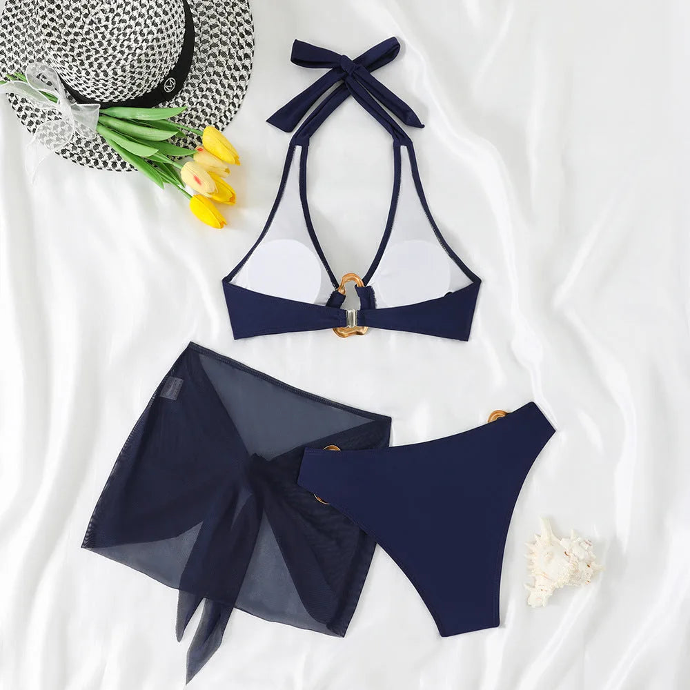 Flat lay of navy halter bikini top and high-leg bottoms with gold rings, shown with a round straw hat and tulips