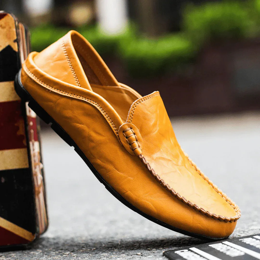 Tan leather loafer balanced on its heel against a vintage flag design box, showing full side profile