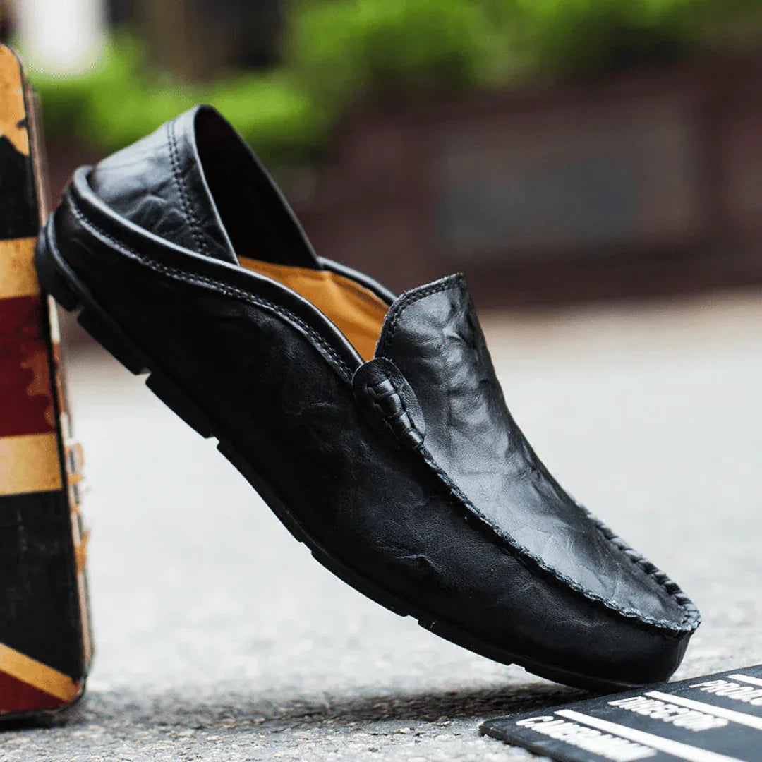 Black leather loafer balanced on its heel beside a flag-patterned box, showing the full silhouette