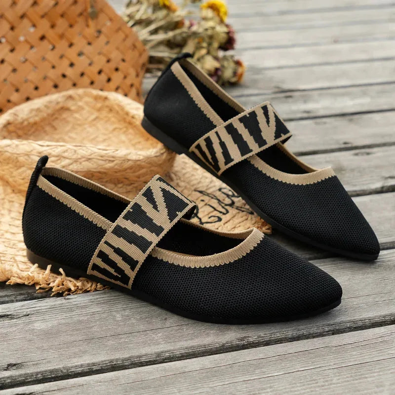Angled view of black knit flats with tan zebra-print strap resting on timber decking beside a straw bag