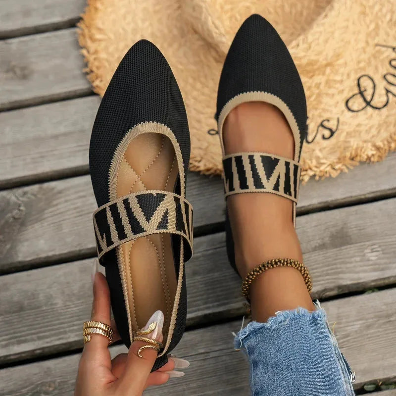 Front angle of black knit pointed-toe flats with gold patterned strap worn on feet, straw hat in background