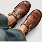 Close-up top view of brown leather T-strap Mary Janes with brogue wingtip detailing, worn with cuffed jeans