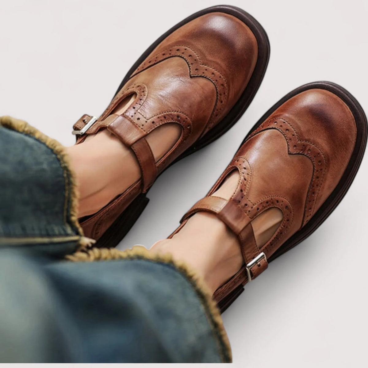 Close-up top view of brown leather T-strap Mary Janes with brogue wingtip detailing, worn with cuffed jeans