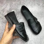 Hand holding a black leather flat beside its matching shoe, showing soft knot detail and square toe on a grey rug