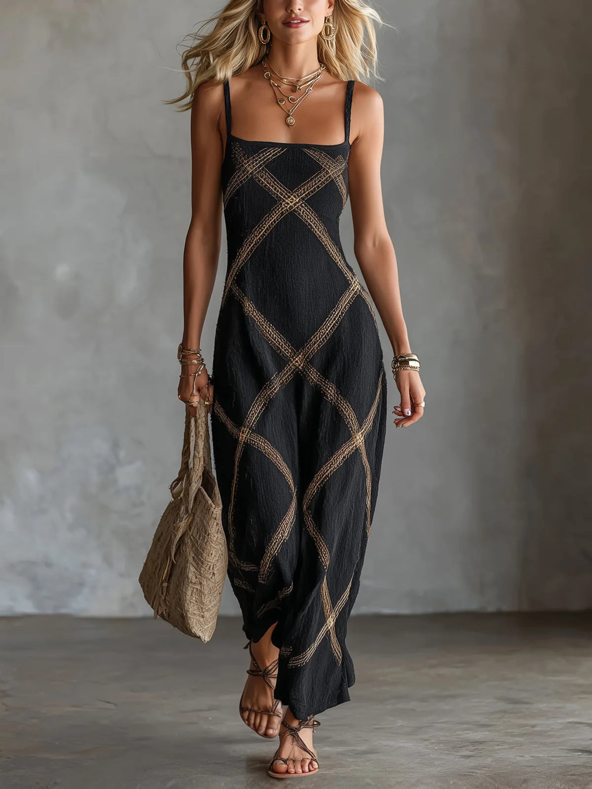 Model wearing a black sleeveless maxi dress with a gold geometric crisscross pattern and square neckline, holding a woven tote