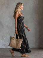 Side profile of model in black sleeveless maxi dress with gold geometric trim, carrying a woven tote bag