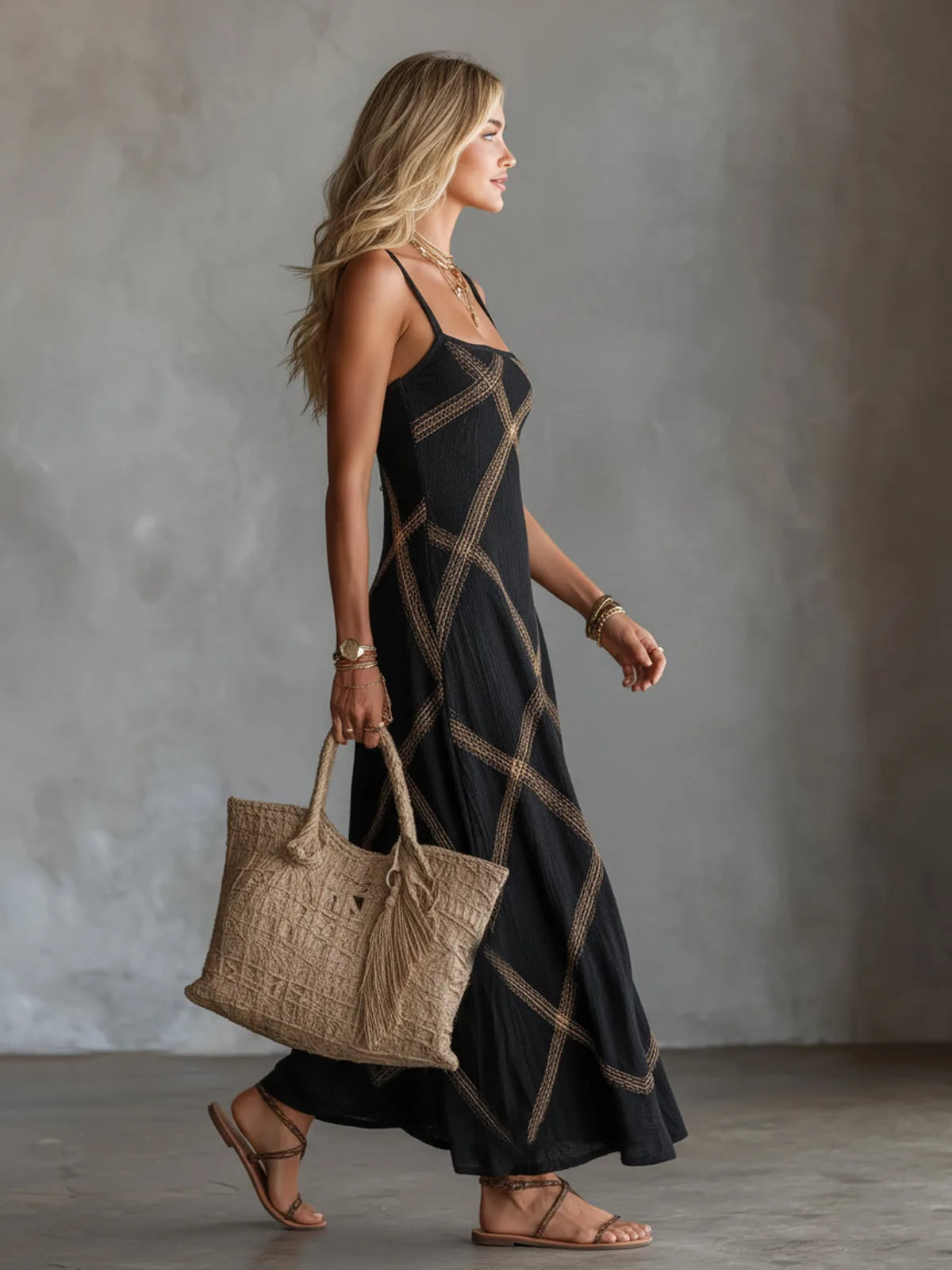 Side profile of model in black sleeveless maxi dress with gold geometric trim, carrying a woven tote bag