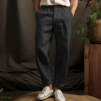 Front view of charcoal black linen tapered trousers with cuffed hems, worn with white sneakers on a woven stool.