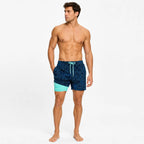 Man standing in navy palm-print swim shorts with mint green compression liner showing at the hem, front view