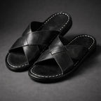 Pair of black crossover strap slide sandals shown from the side against a dark backdrop