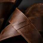 Close-up of the woven crossover strap detail on the brown sandal, showing leather grain and stitching